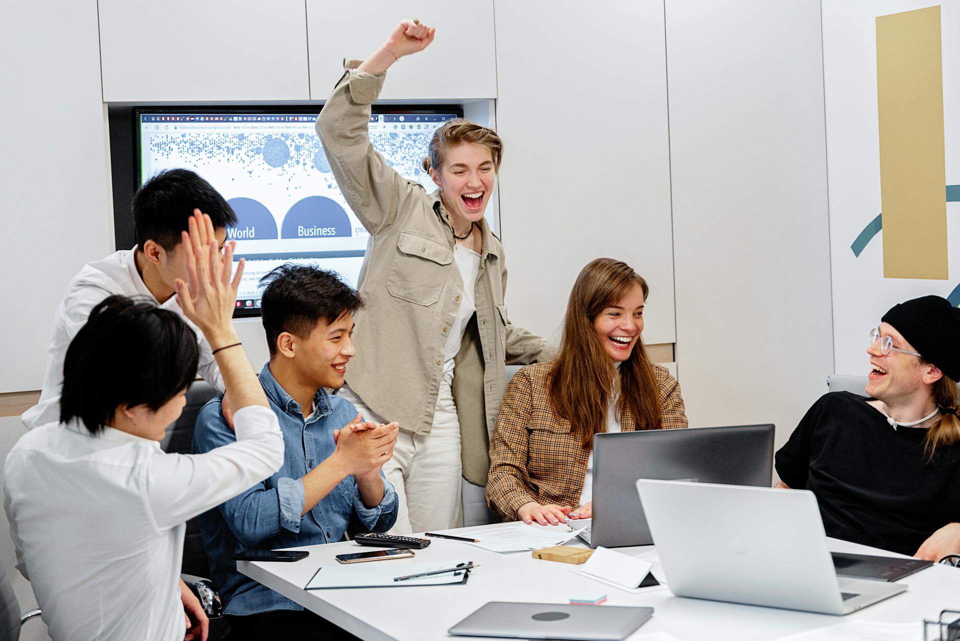 A group of coworkers in a meeting room celebrate around a table with laptops and papers, smiling, clapping, and raising their hands after a successful moment.