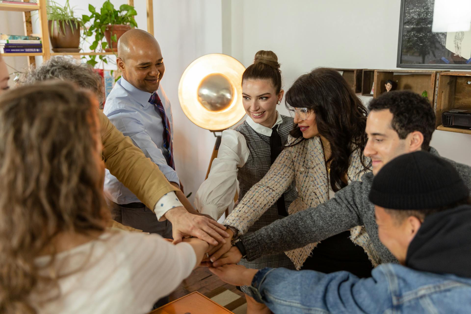 A diverse group of coworkers stand around a table in an office, smiling as they stack their hands together in a team huddle.