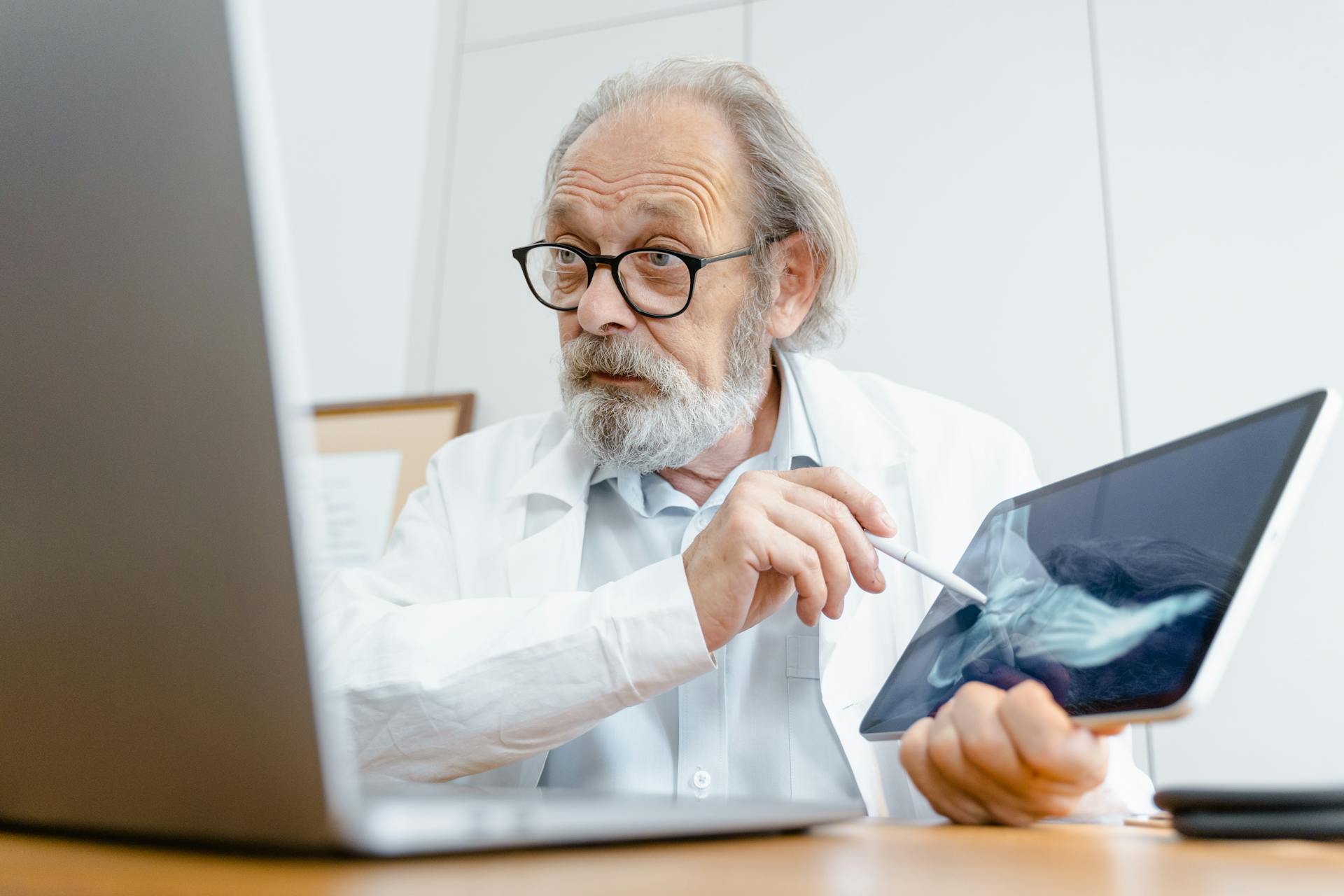 Doctor in a white coat reviews an X-ray on a tablet and points with a stylus while working at a laptop.