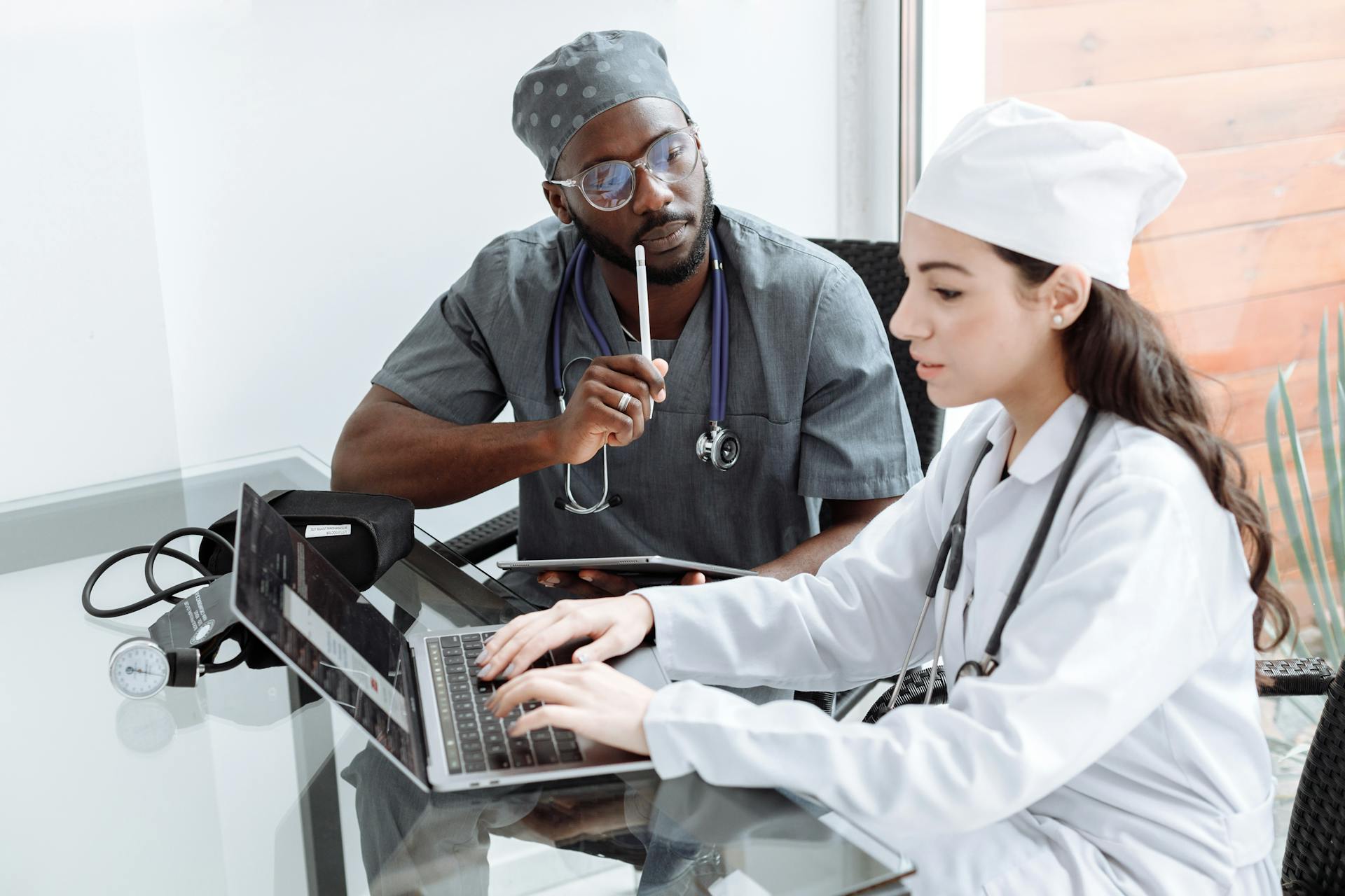 Two healthcare professionals review information on a laptop at a glass table; one types while the other, wearing scrubs and a stethoscope, holds a tablet and stylus.